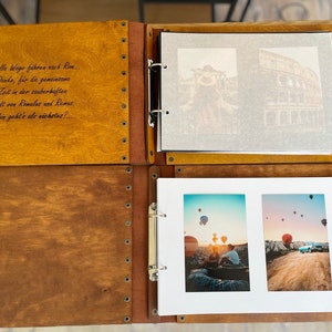 K&ouml;nnte beinhalten: Ein Holzfotoalbum mit braunem Lederbezug und Metallringen. Das Album ist ge&ouml;ffnet und zeigt zwei Seiten mit Fotos vom Kolosseum in Rom und Hei&szlig;luftballons &uuml;ber Kappadokien, T&uuml;rkei. Das Album hat ein Zitat auf Deutsch auf der Vorderseite: "alle Wege f&uuml;hren nach Rom. Danke, f&uuml;r die gemeinsame Zeit in der zauberhaften Stadt von Romales and Remes. Wohin geht's als n&auml;chstes? ..."