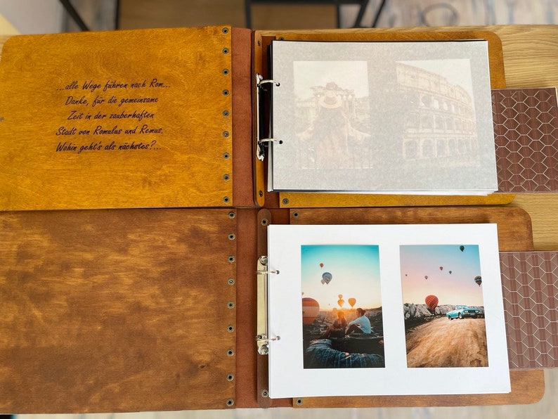 K&ouml;nnte beinhalten: Ein Holzfotoalbum mit braunem Lederbezug und Metallringen. Das Album ist ge&ouml;ffnet und zeigt zwei Seiten mit Fotos vom Kolosseum in Rom und Hei&szlig;luftballons &uuml;ber Kappadokien, T&uuml;rkei. Das Album hat ein Zitat auf Deutsch auf der Vorderseite: "alle Wege f&uuml;hren nach Rom. Danke, f&uuml;r die gemeinsame Zeit in der zauberhaften Stadt von Romales and Remes. Wohin geht's als n&auml;chstes? ..."