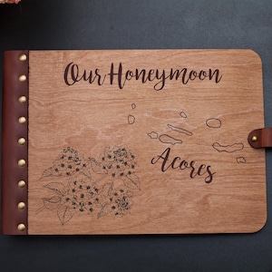 May include: Wooden honeymoon journal with brown leather spine and clasp. The cover is engraved with the words "Our Honeymoon" and "Acores", along with a floral design and a map of the Azores islands.
