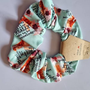 Peut inclure: Un chouchou bleu clair avec un motif répété d'un visage de femme, de fleurs et de texte. Le texte comprend les mots "Serial Killer" et "needs to be acknowledged". Une petite étiquette marron est attachée.