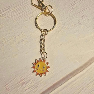 Könnte beinhalten: Ein goldfarbener Schlüsselanhänger mit einem Karabinerhaken, einem Spaltring und einer Kette. Der Anhänger ist eine lächelnde Sonne mit gelbem Zentrum und orangefarbenen Strahlen. Der Schlüsselanhänger liegt auf einer hellen Holzoberfläche.