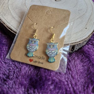 May include: Owl-shaped earrings with gold hooks. The owls are blue, yellow, and pink. The earrings are displayed on a brown card with the words "handmade with love". The background is a purple, textured fabric.