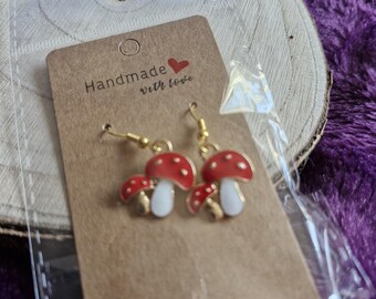 May include: A pair of handmade mushroom earrings with red caps and white spots, and white stems. The earrings are on a brown card that reads "Handmade with love" with a red heart. The earrings are displayed on a wooden slice.