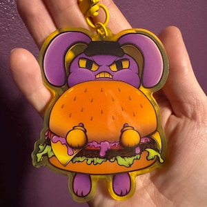 Op de afbeelding: Een heldere acryl sleutelhanger met een cartoon paars konijn karakter met een boze uitdrukking, met een zwarte hoed en een grote hamburger. De sleutelhanger heeft een gele sluiting.