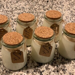 Puede incluir: Seis tarros de vidrio con tapas de corcho y etiquetas de papel marr&oacute;n. Los tarros contienen velas blancas. Las etiquetas tienen texto escrito a mano, incluyendo "Japanese Cherry Blossom", "Rosemary Sage" y "Lavender Sage".