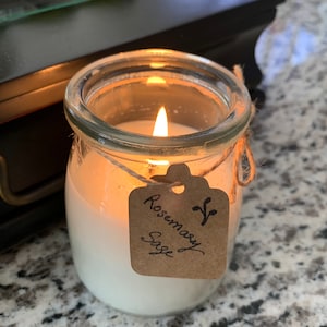 Puede incluir: Una vela en un tarro de vidrio transparente con una vela de cera blanca en su interior. La vela est&aacute; encendida y la llama es visible. El tarro tiene una etiqueta de papel marr&oacute;n atada alrededor con el texto "Rosemary Sage" escrito en tinta negra.