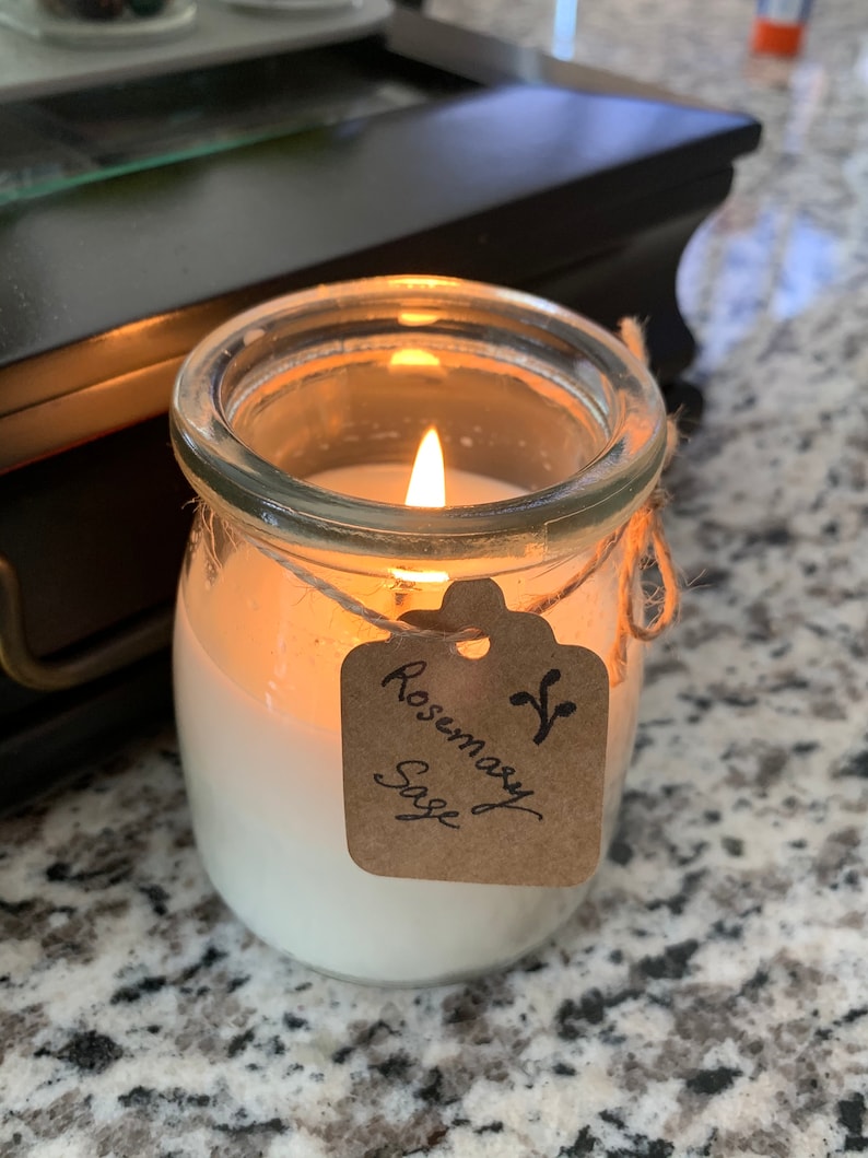 Puede incluir: Una vela en un tarro de vidrio transparente con una vela de cera blanca en su interior. La vela est&aacute; encendida y la llama es visible. El tarro tiene una etiqueta de papel marr&oacute;n atada alrededor con el texto "Rosemary Sage" escrito en tinta negra.