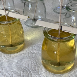 Puede incluir: Primer plano de dos frascos de vidrio llenos de un l&iacute;quido amarillo y una mecha blanca. Los frascos est&aacute;n sobre una superficie blanca con un soporte de mecha de metal.