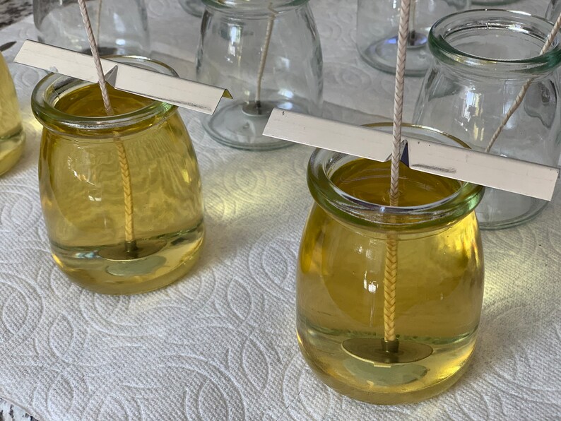 Puede incluir: Primer plano de dos frascos de vidrio llenos de un l&iacute;quido amarillo y una mecha blanca. Los frascos est&aacute;n sobre una superficie blanca con un soporte de mecha de metal.