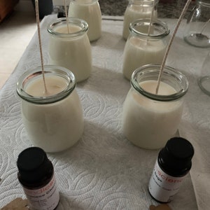 Puede incluir: Seis tarros de vidrio transparentes con cera blanca y mechas, colocados sobre una superficie blanca. Dos peque&ntilde;os frascos de aceite esencial se encuentran en primer plano, uno etiquetado como "Lemongrass Citrus" y el otro como "Sage & Clary Sage".