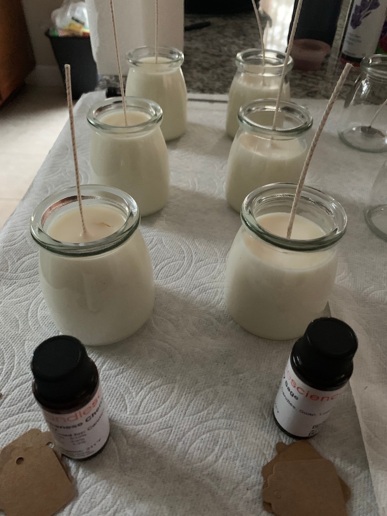 Puede incluir: Seis tarros de vidrio transparentes con cera blanca y mechas, colocados sobre una superficie blanca. Dos peque&ntilde;os frascos de aceite esencial se encuentran en primer plano, uno etiquetado como "Lemongrass Citrus" y el otro como "Sage & Clary Sage".