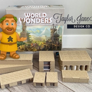 Op de afbeelding: Een bordspel genaamd "World Wonders" met een stripfiguur met een rol en een standbeeld. De set bevat beige bouwcomponenten en een doos met de speltitel en artwork. Het Taylor Jane Design Co. logo is ook zichtbaar.