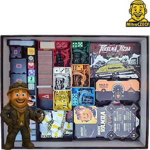 Op de afbeelding: Een bordspelbox gevuld met spelonderdelen, waaronder kaarten, dobbelstenen en miniatuurvoertuigen. De doos toont de tekst "TEKELNA JIZDA" en een stripfiguur met een stuurwiel. De spelstukken zijn in gekleurde compartimenten geplaatst.