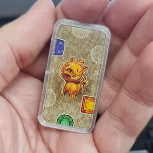 Può includere: Una carta da gioco rettangolare con un personaggio di maiale dei cartoni animati con un'espressione amichevole. La carta ha uno sfondo beige con gemme colorate e un motivo a pois. La carta è tenuta in mano da una persona.