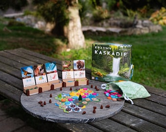 Puede incluir: Un juego de mesa titulado Krajiny Kaskádie que incluye una caja con la ilustración de una cascada, soportes de cartón para cartas con animales, un tablero hexagonal central, fichas de piñas marrones y una bolsa de tela verde claro.