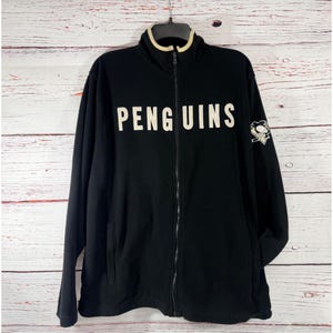 Puede incluir: Chaqueta negra con cremallera con la palabra "PENGUINS" en letras blancas en el pecho. La chaqueta tiene un cuello color crema y un parche con el logotipo del equipo en la manga izquierda. Con dos bolsillos delanteros.