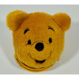 Puede incluir: Una cara de Winnie the Pooh amarilla de felpa, con ojos, nariz y boca sonriente negros. Tiene un asa amarilla, posiblemente un monedero o una bolsa pequeña. El diseño es un personaje de dibujos animados clásico.
