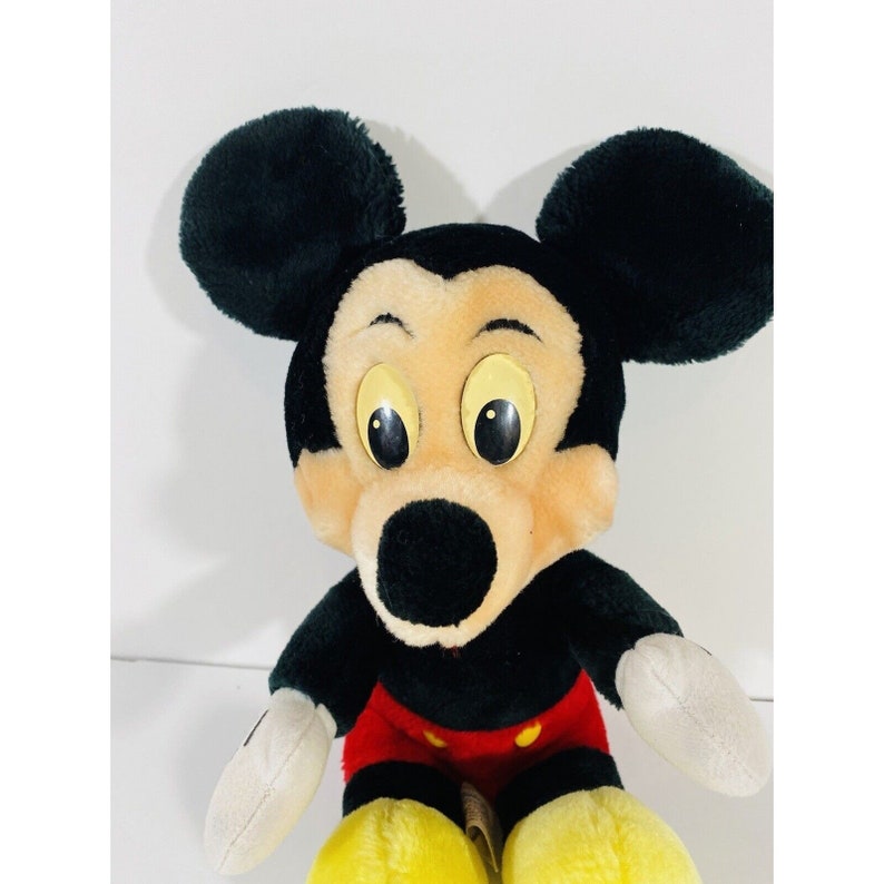 Puede incluir: Un juguete de peluche de Mickey Mouse con orejas negras, nariz negra y ojos amarillos. El juguete lleva pantalones cortos rojos con botones blancos y zapatos amarillos.