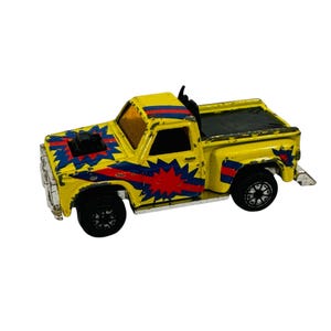 May include: A vintage yellow toy pickup truck with black wheels and a black bed cover. The truck features red and blue starburst and stripe designs on the hood and doors. The toy has a black exhaust pipe and a silver front bumper.