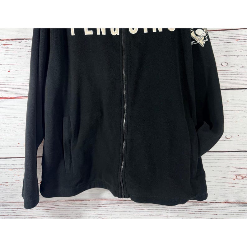Puede incluir: Chaqueta negra con cremallera con la palabra "Penguins" en letras blancas en la parte superior. Un logotipo del equipo est&aacute; en la manga superior derecha. La chaqueta tiene dos bolsillos delanteros y una cremallera completa.