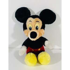 Puede incluir: Un juguete de peluche de Mickey Mouse con pelaje negro, ojos grandes amarillos y un atuendo rojo y amarillo.