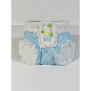 Puede incluir: Un macetero de cerámica azul y blanco con forma de mono de bebé con ribete rosa y blanco y detalles florales.