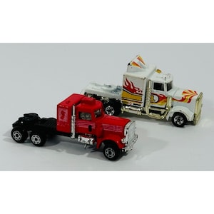 May include: Two toy semi-trucks. One is red with a black trailer and the other is white with flame decals. Both have black wheels and silver accents. The trucks are on a white background.