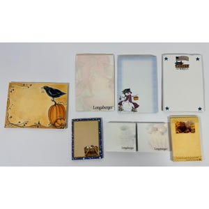 May include: A collection of decorative notepads with various seasonal designs. One notepad features a crow perched on a pumpkin. Other designs include a snowman, a basket of eggs, and a patriotic theme with stars and stripes.