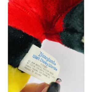 Puede incluir: Una etiqueta blanca con texto negro que dice "Disneyland Walt Disney World" e incluye informaci&oacute;n sobre los materiales y el origen del producto. La etiqueta est&aacute; unida a un juguete de peluche con piel roja, negra y amarilla.
