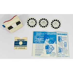 Puede incluir: Un visor View-Master vintage con tres carretes y materiales impresos relacionados. El visor es de color blanco roto con detalles en rojo. Los carretes son en blanco y negro. Los materiales impresos incluyen un formulario de pedido por correo y un folleto con texto e ilustraciones azules.