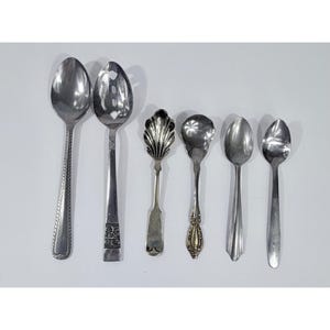 May include: A collection of six silver spoons of varying designs. The spoons include a slotted spoon, a spoon with a shell-shaped bowl, and spoons with decorative handles. The spoons are arranged on a white surface.
