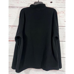 Puede incluir: Un jersey de forro polar negro con cuello alto. La camisa de manga larga est&aacute; colgada de una percha negra sobre un fondo de tablones de madera blancos y marrones. La prenda est&aacute; hecha de un material suave y c&aacute;lido.