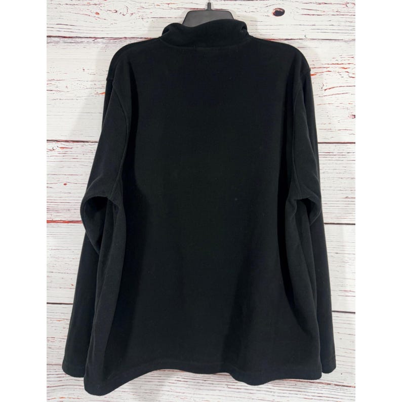 Puede incluir: Un jersey de forro polar negro con cuello alto. La camisa de manga larga est&aacute; colgada de una percha negra sobre un fondo de tablones de madera blancos y marrones. La prenda est&aacute; hecha de un material suave y c&aacute;lido.