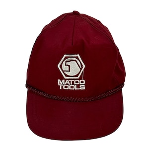 Può includere: Cappellino da baseball bordeaux con logo ricamato bianco e scritta "MATCO TOOLS". Il cappellino ha una visiera curva e un dettaglio in corda intrecciata lungo il bordo della visiera.