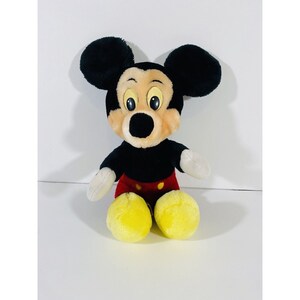 Puede incluir: Un juguete de peluche Mickey Mouse negro con zapatos amarillos y pantalones cortos rojos. El juguete tiene una nariz negra y ojos grandes y redondos.