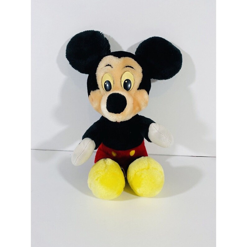Puede incluir: Un juguete de peluche Mickey Mouse negro con zapatos amarillos y pantalones cortos rojos. El juguete tiene una nariz negra y ojos grandes y redondos.