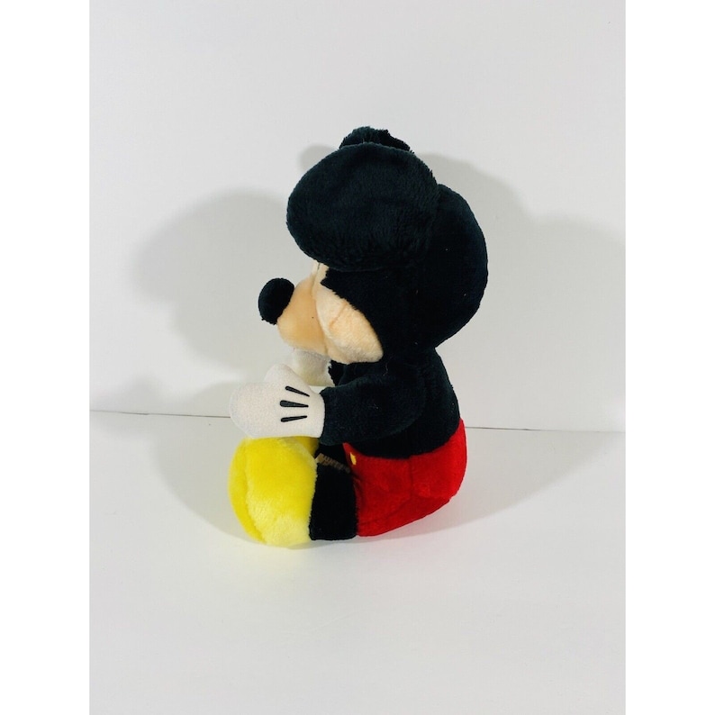 Puede incluir: Un juguete de peluche de Mickey Mouse sentado con las piernas cruzadas. Lleva pantalones cortos rojos y zapatos amarillos. Sus orejas son negras y su cara es de color marr&oacute;n claro.