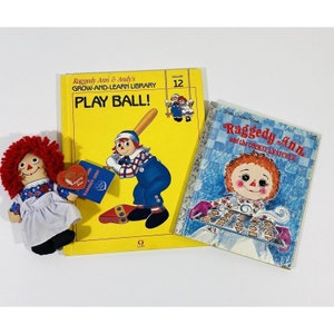 May include: Two Raggedy Ann children's books. The first book is yellow with the title "Play Ball!" and features Raggedy Ann playing baseball. The second book is blue with the title "Raggedy Ann and the Cookie Snatcher" and features Raggedy Ann holding a tray of cookies.