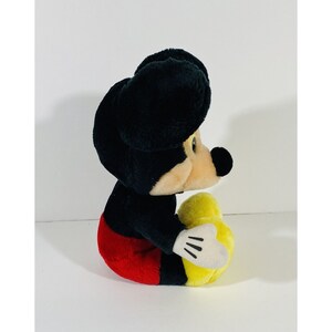 Puede incluir: Un juguete de peluche de Mickey Mouse sentado con las piernas cruzadas. Lleva pantalones cortos rojos y zapatos amarillos. Sus orejas son negras y su cara es de color marr&oacute;n claro.