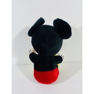 Puede incluir: Un juguete de peluche Mickey Mouse negro y rojo sentado en una superficie blanca. El juguete tiene grandes orejas negras y lleva pantalones cortos rojos.