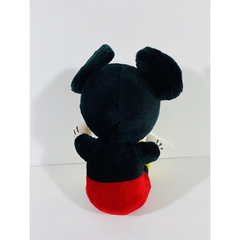 Puede incluir: Un juguete de peluche Mickey Mouse negro y rojo sentado en una superficie blanca. El juguete tiene grandes orejas negras y lleva pantalones cortos rojos.