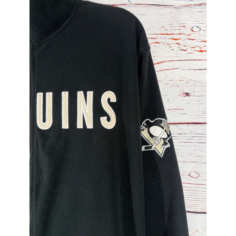 Puede incluir: Chaqueta negra de forro polar con cremallera y la palabra "UINS" en letras color crema. La manga derecha tiene un parche con el logotipo del equipo con un ping&uuml;ino y palos de hockey.