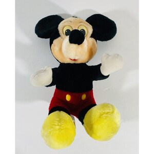 Puede incluir: Un juguete de peluche de Mickey Mouse con pelaje negro, pantalones cortos rojos y zapatos amarillos. El juguete tiene una expresi&oacute;n amigable con ojos grandes y una amplia sonrisa.