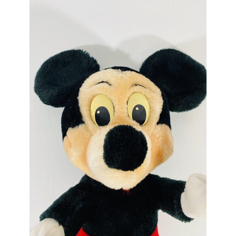 Puede incluir: Un juguete de peluche de Mickey Mouse con orejas negras, nariz negra y grandes ojos amarillos. El juguete lleva un atuendo rojo y negro.