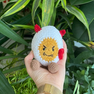 May include: A white crocheted egg-shaped toy with a yellow face and red horns. The toy has a grumpy expression and is being held by a hand wearing a gold watch.