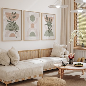 May include: A light beige sofa with a wooden frame and three pillows. The sofa is in a living room with a coffee table, a woven ottoman, and three framed abstract art prints with green and pink geometric shapes and leaves.