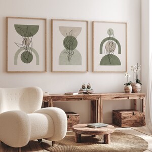 May include: Three framed abstract art prints with a minimalist design featuring green and beige geometric shapes and line drawings of leaves. The prints are hanging on a white wall above a wooden table with a white armchair and a small wooden table with a book on top.
