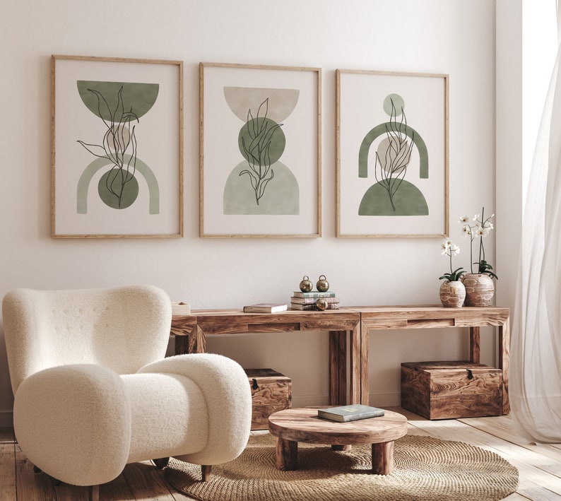 May include: Three framed abstract art prints with a minimalist design featuring green and beige geometric shapes and line drawings of leaves. The prints are hanging on a white wall above a wooden table with a white armchair and a small wooden table with a book on top.