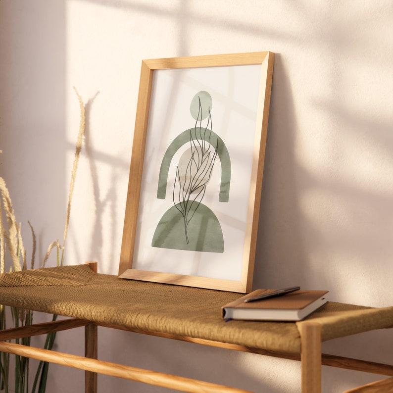 May include: A framed print with a minimalist design featuring a green leaf and abstract shapes in shades of green and beige. The print is hanging on a wall above a wooden bench.