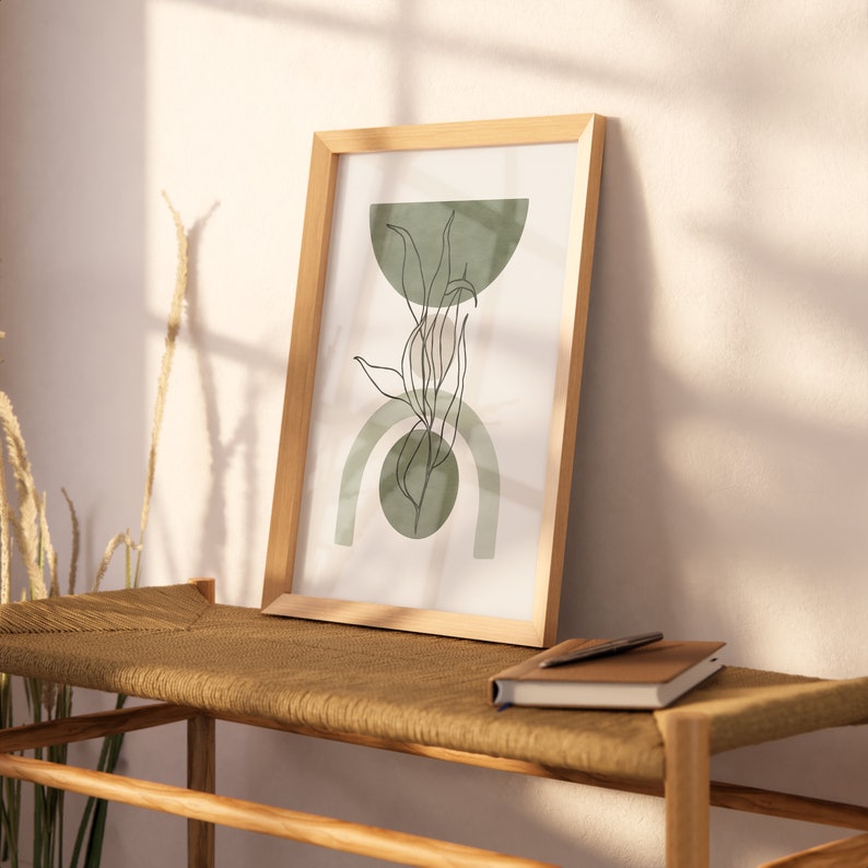 May include: A framed print with a minimalist design featuring a green leaf and geometric shapes in shades of green. The print is hanging on a light wood wall shelf with a brown woven seat.